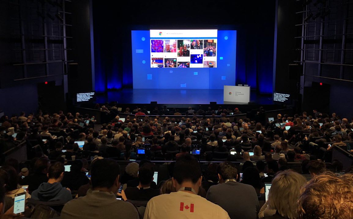 A large audience in a dark auditorium watches a conference presentation with a social media wall displayed on a screen above a stage with a podium.