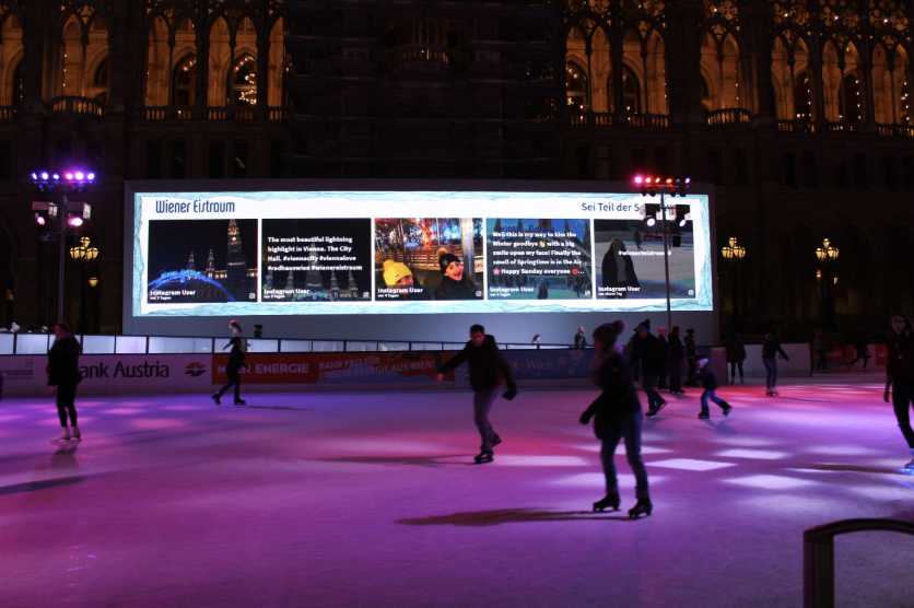 An outdoor ice skating rink at night features a giant screen displaying a Walls.io social media wall with Instagram posts from visitors at Wiener Eistraum in front of the illuminated Vienna City Hall. Skaters enjoy the rink while user-generated social media content lights up the backdrop.