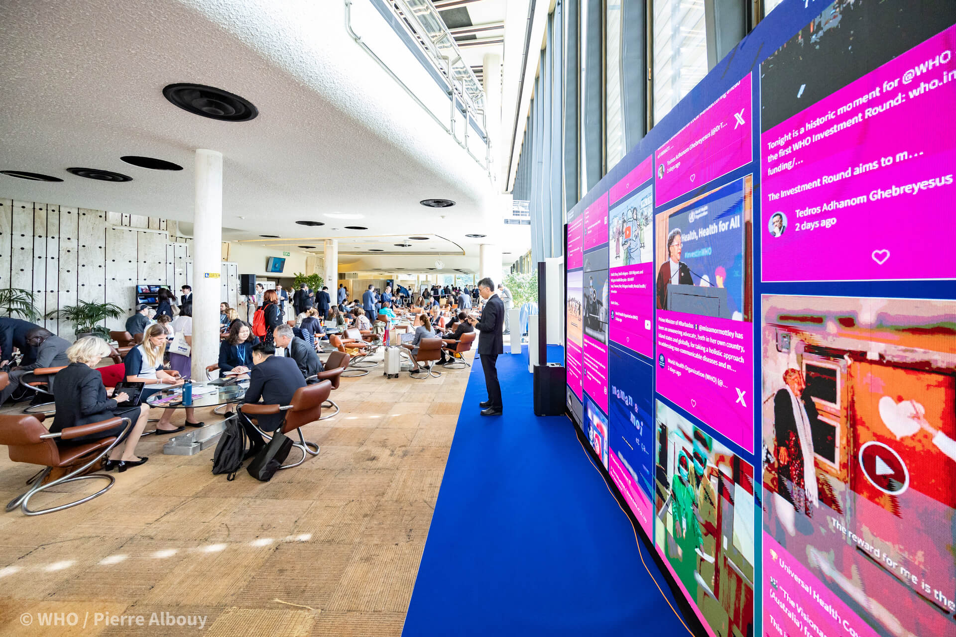 A large social wall audience activation platform for events displayed in the lobby of a WHO event, showing live posts from X and other platforms on a floor-to-ceiling LED screen while attendees work nearby.