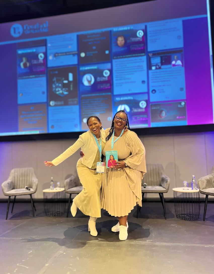 Two attendees posing and smiling on stage at a Festival, with a social wall for conferences displaying live event posts on the main screen behind them.