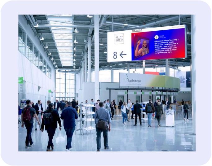 Trade show hallway at Koelnmesse with an audience activation platform on a hanging digital sign, displaying an attendee's social media post to passing visitors.